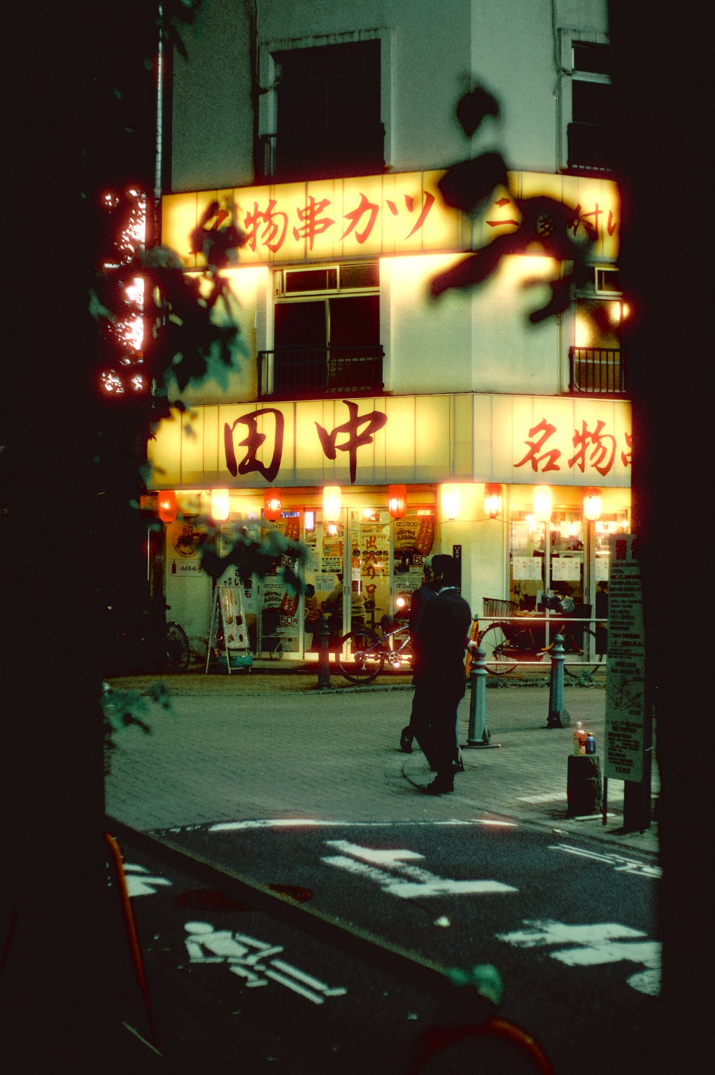 A warmly lit neighborhood restaurant seen through dark foliage.